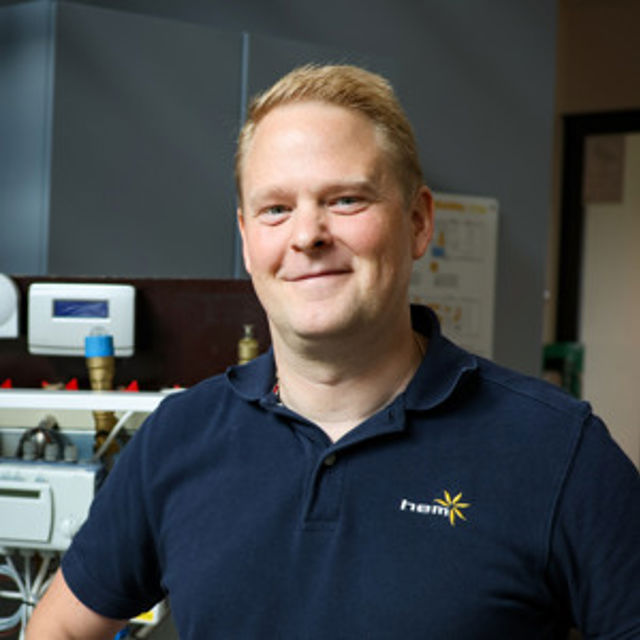 Leende person i mörkblå pikétröja med HEM-logga på bröstet, står inomhus i en teknisk miljö med utrustning och instrument i bakgrunden.