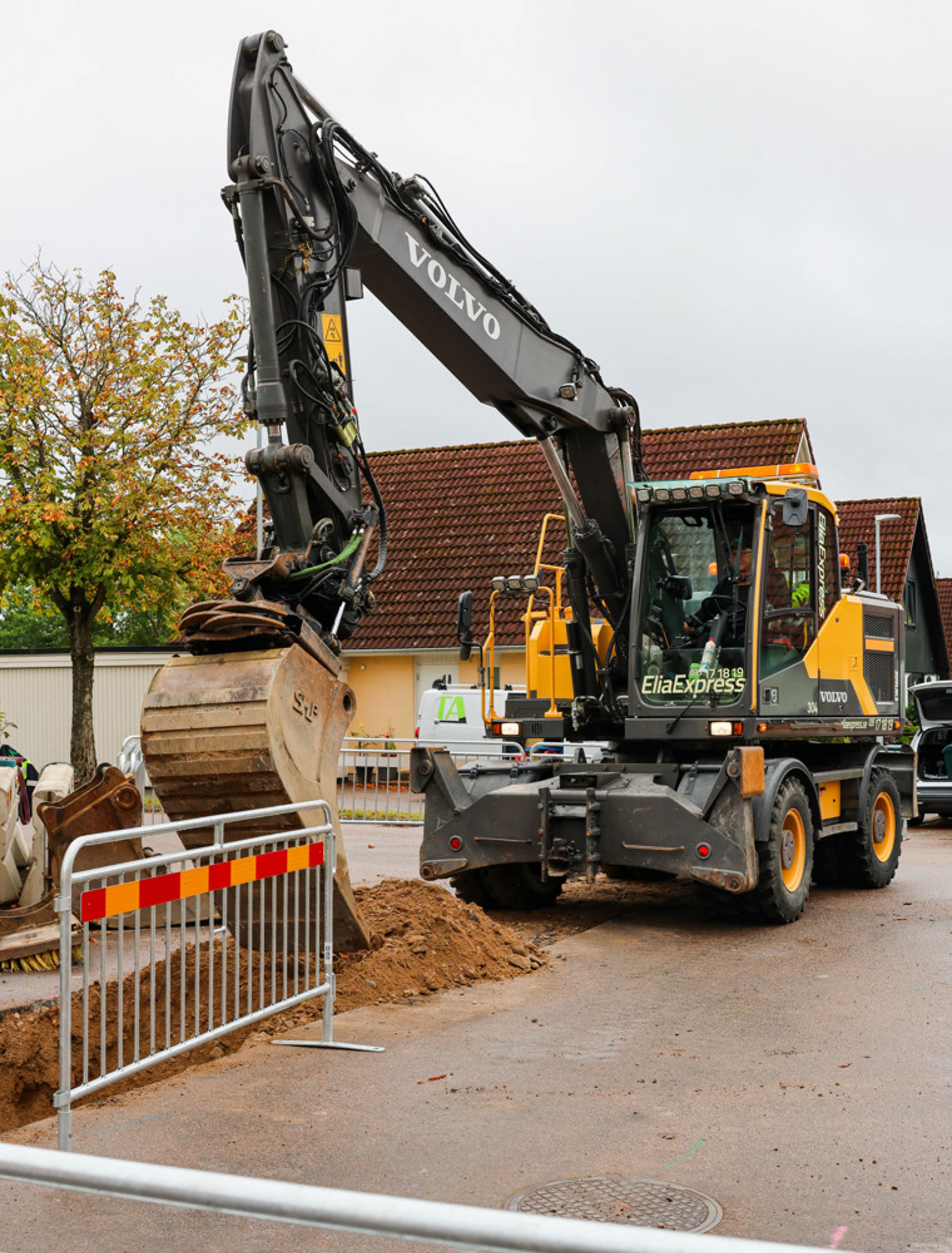 Grävarbete längs en gata – synliga rör och ledningar i marken, jordmassor runt omkring, arbetszon insatt med avspärrningar, bostadshus och vegetation i bakgrunden